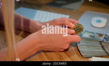 Crop view of hands of woman holding shiny metal bitcoin sitting at wooden desk with wad of cash nearby at night Stock Photo