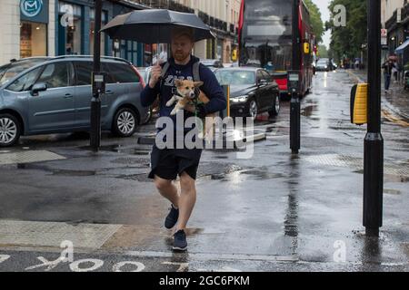 06/08/2021. London, UK. A man carries his dog as he shelter sunder an umbrella during a rain shower in Greenwich, South East London. Photo credit: Geo Stock Photo