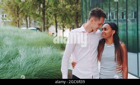 Tall young man kissing her african girlfriend while walking down the street. She telling him about something. Relationship concept Stock Photo