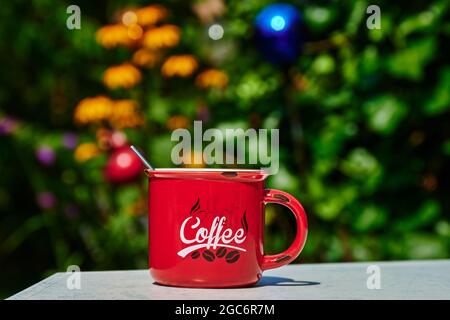 A red coffee cup on a garden table with blurred flowers in the background. Stock Photo