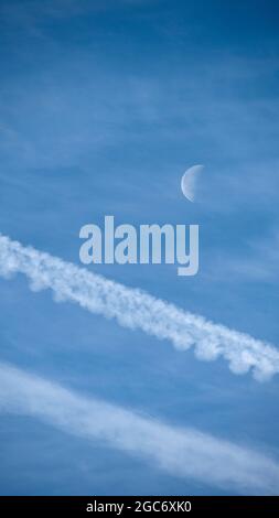 Airplane contrails and waning moon against blue sky Stock Photo