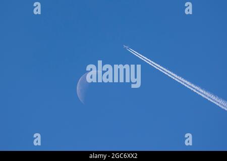 Airplane leaving contrails against blue sky with waning moon Stock Photo