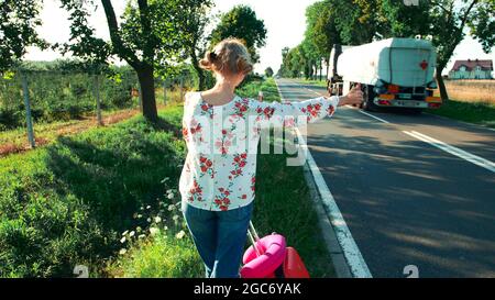 Traveler woman hitchhiking on a sunny road and walking. Young happy backpacker woman looking for a ride to start a journey on a sunlit country road. Stock Photo