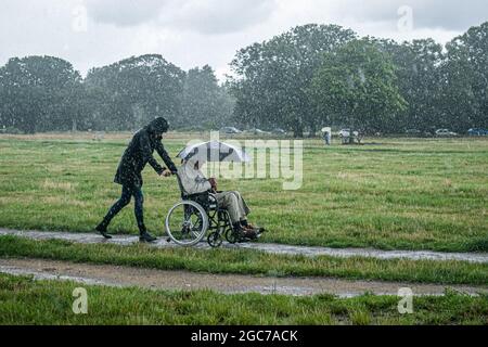 London, UK. 07th Aug, 2021. WIMBLEDON LONDON 7 August  2021. A woman pushing a man in a wheelchair holding an umbrella during  a downpour on Wimbledon Common. The met office has issued thunderstorm warning with flash flooding over parts of the United Kingdom  that is likely to cause travel disruptions. Credit amer ghazzal/Alamy Live News. Credit: amer ghazzal/Alamy Live News Stock Photo