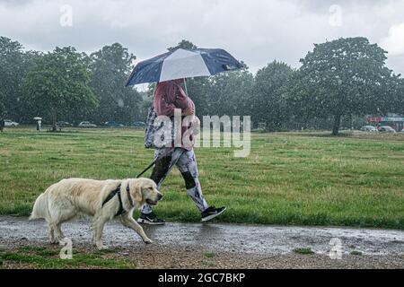 London, UK. 07th Aug, 2021. WIMBLEDON LONDON 7 August  2021. A woman  with an umbrella walks her dog  during  a  heavy downpour on Wimbledon Common. The met office has issued thunderstorm warning with flash flooding over parts of the United Kingdom  that is likely to cause travel disruptions. Credit amer ghazzal/Alamy Live News. Credit: amer ghazzal/Alamy Live News Stock Photo