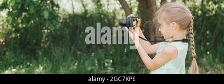 a happy little seven year old kid girl photographs a summer natural landscape with a camera using live view. children adopt their parents hobbies. sum Stock Photo