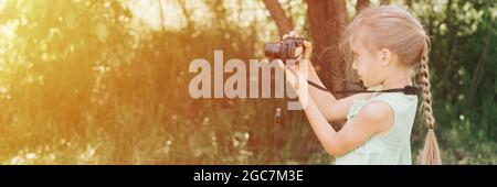 a happy little seven year old kid girl photographs a summer natural landscape with a camera using live view. children adopt their parents hobbies. sum Stock Photo