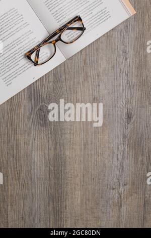 a pair of reading glasses sitting on top of an open book on a wooden table, viewed from above. Stock Photo