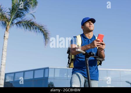 African delivery man browsing address on mobile phone map gps app for fast food order at electric scooter - Focus on courier man face Stock Photo