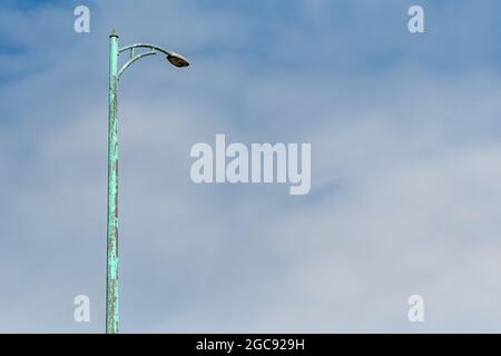 Looking up at a tall light green metal street light against a sky with thin clouds. Paint on the pole is faded green buy much paint has fallen off. Stock Photo