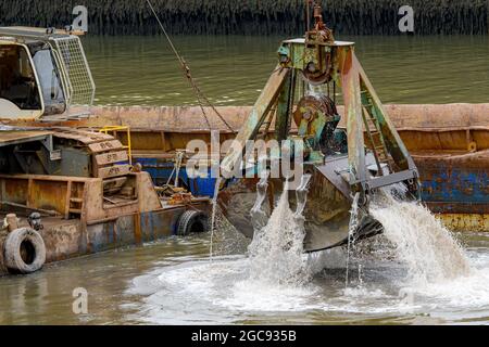 Closeup view of a large dredge scoop rising from the harbor. Water flows from it. Dredge crane to the left (operator hidden), barge behind. Stock Photo