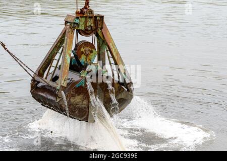 Closeup view of a large dredge rising from the water in a harbor. Water flows from the closed scoop. Only scoop and water visible in scene. Stock Photo