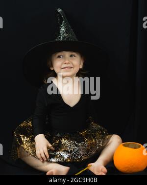 A little cheerful girl in a witch costume-a hat and a skirt with a spider web-sits on a black background and looks away. Next to it is a bucket-pumpki Stock Photo