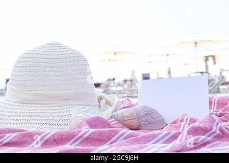 Summer vacation still life. White straw hat, seashell on red and white towel.Beach umbrellas in the background. Blank invitation card or greeting card Stock Photo