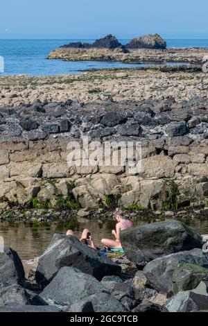 young couple sitting on rocks, young couple addicted to phones, couple looking at smartphones, young couple sitting on rocks at seasdie, using devices Stock Photo