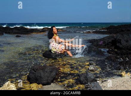 Older Hawaiian woman wades into the shallows and is sitting on a black lava rock kicking and splashing. Stock Photo