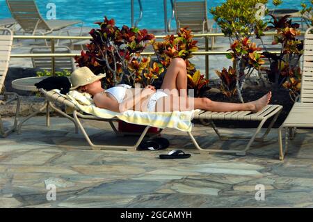 Fresh Hawaiian air on the Big Island, has lulled this female to sleep besides the swimming pool.  She is wearing a white bikini and a straw hat over h Stock Photo