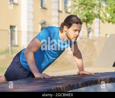 Sportive guy training arms while doing push ups on bench in park on summer day Stock Photo