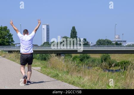 Close up of a senior man jogging away from the camera making a V-sign along a footpath alongside a canal on a sunny summer day Stock Photo