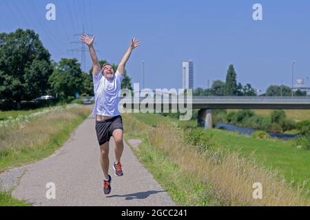 Joyful exuberant middle-aged man out jogging along a river celebrating his good health and sunny weather raising his arms in the cheering Stock Photo