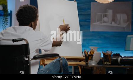 African american disabled young artist drawing vase on canvas and easel while sitting in wheelchair at workshop studio. Black paralyzed person with disability working on fine art concept Stock Photo