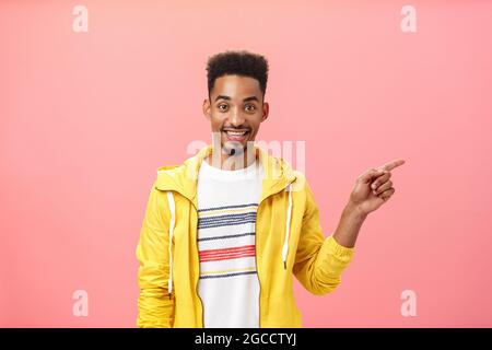 Waist-up shot of friendly relaxed good-looking dark skinned guy with beard in yellow jacket pointing right and smiling joyfully at camera sharing Stock Photo