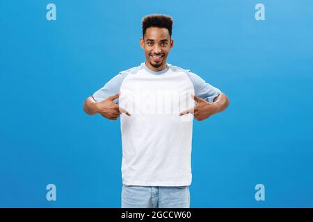Pleased and delighted good-looking man showing cool t-shirt pointing at himself with proud satisfied expression standing over blue background happy Stock Photo