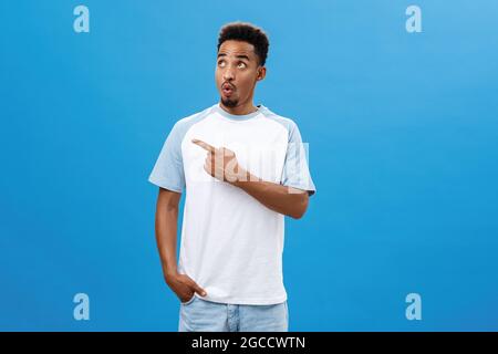 Indoor shot of amazed and thrilled amused handsome dark-skinned guy with beard in casual t-shirt pointing and gazing left with folded lips from Stock Photo