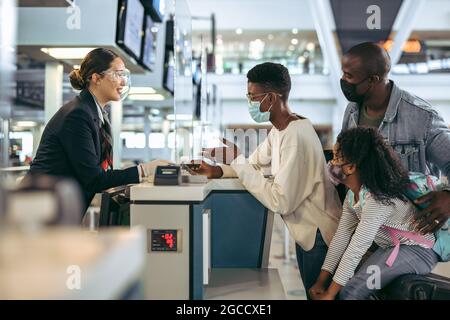 Passengers standing at check-in counter with airport staff during pandemic. African family in covid-19 outbreak at check-in desk of airport. Stock Photo