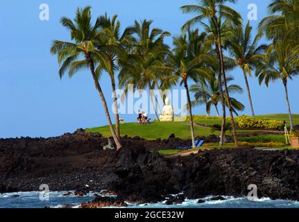 Couple relax on a bench beneath palm trees at a resort on the Big Island of Hawaii.  Ocean waves splash against the lava rock beach. Stock Photo