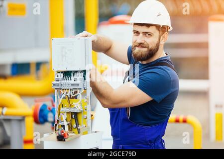 Electrician in a helmet and overalls works adjusts the equipment looks into the camera and smiles. A positive portrait of a kind Caucasian worker with a beard at work. Energy Industry Service. Advertising photo for the site. Stock Photo