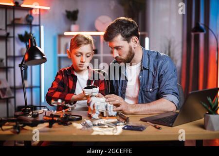 Caring father controlling repairing process of toy robot dog by his little son. Bearded man teaching boy fixing broken appliances at home. Educational moments during parenthood. Stock Photo