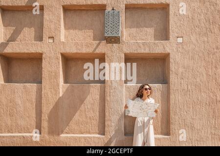 Happy woman travels alone and looks at a map against the wall of an ancient city in the Middle East region or somewhere in Morocco. Stock Photo