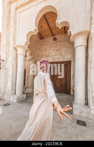 Woman traveler wearing red turban and biege long dress making follow me pose at the old narrow streets of Bur Dubai and Creek. Travel culture and sigh Stock Photo