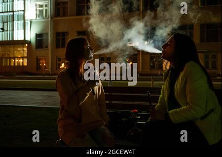Two girlfriends smoke on the background of the night city. Women smoke cigarettes and vape late at night. Bad habit. Stock Photo