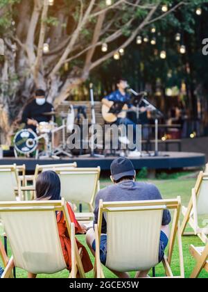 Back view of Asian young couple sit to relax on white deck chairs with tables for dinner in lawn is surrounded by shady green grass with blurred image Stock Photo