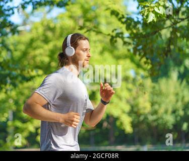 Young male sportsman wearing headphones jogging in green park in morning Stock Photo