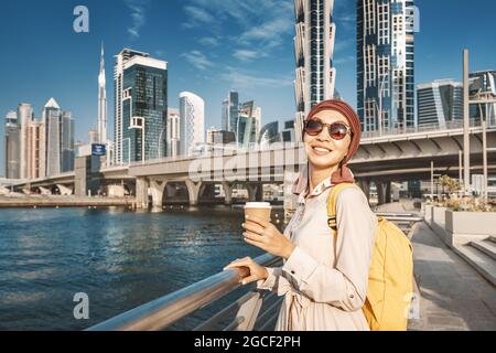 Happy muslim woman with yellow backpack walks along the Dubai Canal embankment and drinks delicious coffee from a paper cup. Tourism and businesswoman Stock Photo