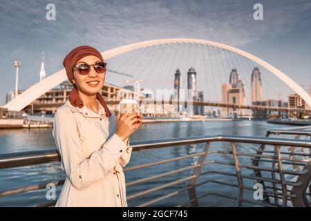 Happy muslim woman with yellow backpack walks along the Dubai Canal embankment and drinks delicious coffee from a paper cup. Tourism and businesswoman Stock Photo