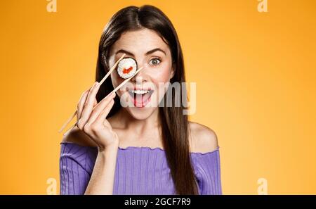 Excited girl holding sushi with salmon and rise near eye, eating asian food with chopsticks and smiling amazed at camera, orange background Stock Photo