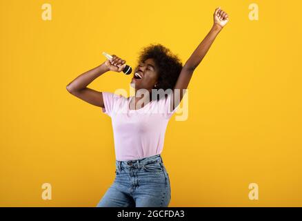 Music lover. Excited black woman singing songs in microphone and dancing, having fun and enjoying the moment, standing over yellow background, studio Stock Photo