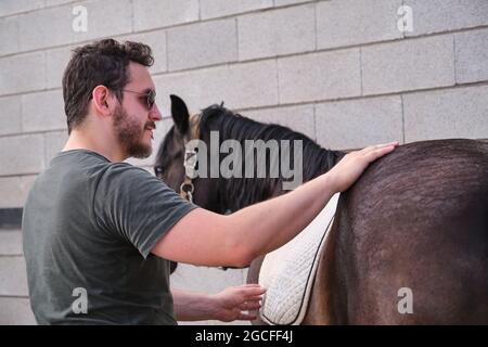 Young latin man putting on the saddle blanket to his horse and stroking it. Stock Photo