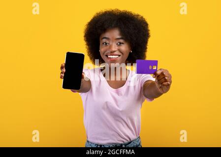 Banking, purchasing concept. Excited black woman showing blank smartphone screen and credit card over yellow studio background, mockup for design. Mob Stock Photo