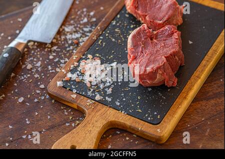 Raw Hereford grass fed beef tenderloin on black slade board with salt, knife and a cup of rocket salad Stock Photo