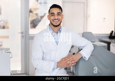 Portrait Of Handsome Arab Stomatologist Doctor Posing In Dental Clinic Interior, Smiling Middle Eastern Dentist In Uniform Standing At Workplace And L Stock Photo