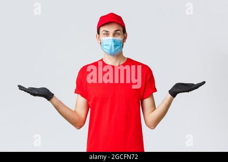 Groceries and packages delivery, covid-19, quarantine and shopping concept. Friendly delivery man in red uniform, face mask and gloves, holding two Stock Photo