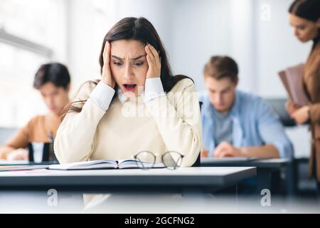 Portrait of shocked female student sitting at desk in classroom, grabbing her head looking at exam questions or results. Worried youth unprepared for Stock Photo