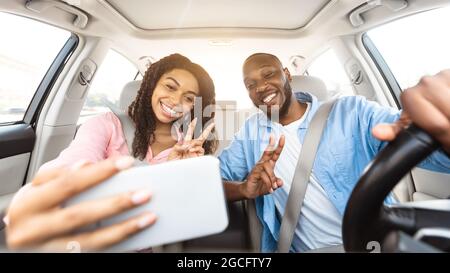 Beautiful young African American couple taking selfie inside car, using mobile phone, showing v victory peace sign gesture. Happy black man and woman Stock Photo