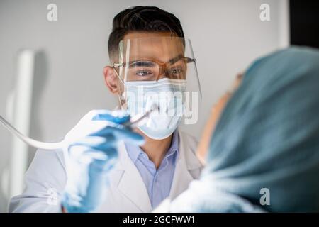 Closeup Shot Of Arab Dentist Doctor In Medical Mask And Face Shield Checking Patient's Teeth, Professional Stomatologist Making Dental Treatment To Mu Stock Photo