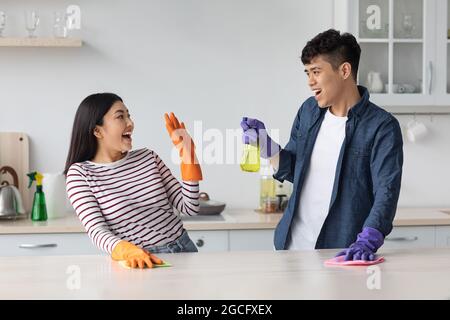 Joyful asian lovers young man and woman in casual clothes having fun while cleaning kitchen together, splashing at each other with cleaning sprays, we Stock Photo